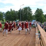 Vairāki dejotāji tradicionālos sarkanos tērpos dejo uz gaiša koka tilta. Liels pūlis cilvēku vēro priekšnesumu, un fonā redzami zaļi koki zem mākoņainām debesīm.