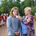 Sieviete ar īsiem blondiem matiem, tērpusies ziedainā blūzē, intervē jaunu vīrieti ar gariem blondiem matiem, turot viņam priekšā mikrofonu. Abi stāv uz koka tilta, aiz viņiem rindā stāv jaunieši vēsturiskos sarkanos tērpos. Fonā redzami zaļi koki.
