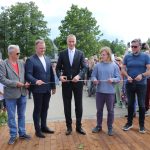 Pieci vīrieši uz koka platformas griež baltu lenti, atzīmējot atklāšanas ceremoniju. Centrālais vīrietis tumšā uzvalkā izmanto zelta šķēres. Apkārt ir vērotāju pūlis un zaļi koki fonā. Pie ziedu groza labajā pusē ir zīme ar tekstu.