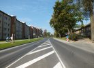 Gara, asfaltēta iela ar baltām ceļa marķējuma līnijām saulainā dienā. Ielas kreisajā pusē redzama rinda ar gaišām daudzdzīvokļu mājām, gar kurām uz ietves staigā cilvēks ar ziliem bērnu ratiņiem. Labajā pusē — vecas koka ēkas un lieli koki. Pie ceļa redzamas zīmes "Stāvēšana aizliegta", "Obligāts virziens pa labi" un "Bīstams līkums pa kreisi, tad pa labi".