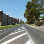 Gara, asfaltēta iela ar baltām ceļa marķējuma līnijām saulainā dienā. Ielas kreisajā pusē redzama rinda ar gaišām daudzdzīvokļu mājām, gar kurām uz ietves staigā cilvēks ar ziliem bērnu ratiņiem. Labajā pusē — vecas koka ēkas un lieli koki. Pie ceļa redzamas zīmes "Stāvēšana aizliegta", "Obligāts virziens pa labi" un "Bīstams līkums pa kreisi, tad pa labi".