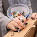Cilvēka roka ievieto dažāda veida izlietotas baterijas kartona savākšanas kastē. Fonā redzams caurspīdīgs spainis, pilns ar citām izlietotām baterijām.