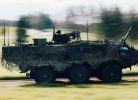 Bruņots militārais transportlīdzeklis, kas pārklāts ar maskēšanās tīklu, ātri brauc pa ceļu, ar kustības izplūdušu zaļu pļavu un kokiem fonā. Uz transportlīdzekļa redzams ložmetējs, pie kura atrodas karavīrs, un plīvo Latvijas karogs.