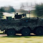 Bruņots militārais transportlīdzeklis, kas pārklāts ar maskēšanās tīklu, ātri brauc pa ceļu, ar kustības izplūdušu zaļu pļavu un kokiem fonā. Uz transportlīdzekļa redzams ložmetējs, pie kura atrodas karavīrs, un plīvo Latvijas karogs.