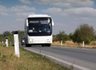 Balts autobuss un aiz tā automašīna brauc pa asfaltētu ceļu ar baltām līnijām. Ceļmalā zāle un balti stabiņi, fonā koki un mākoņainas debesis.