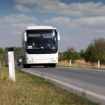 Balts autobuss un aiz tā automašīna brauc pa asfaltētu ceļu ar baltām līnijām. Ceļmalā zāle un balti stabiņi, fonā koki un mākoņainas debesis.
