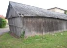 Noliecies vecs koka šķūnis ar tumšiem dēļiem un pelēku, sūnām klātu gofrētu jumtu. Apkārt šķūnim ir zāle, bet fonā redzamas citas ēkas un celiņš.