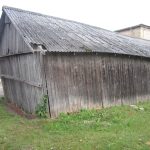 Noliecies vecs koka šķūnis ar tumšiem dēļiem un pelēku, sūnām klātu gofrētu jumtu. Apkārt šķūnim ir zāle, bet fonā redzamas citas ēkas un celiņš.