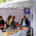 Balta telts ar Luta grāmatu stendu, kurā sievietes aplūko grāmatas koka kastēs uz galdiem. Labajā pusē sieviete baltā t-kreklā ar uzrakstu "Luto.lv" un melnā žaketē stāv pie stenda, smaidot un skatoties pa kreisi. Uz purpursarkaniem baneriem redzams uzraksts "Luta", "Pievienojies un atbalsti" un "Laiks lasīt".