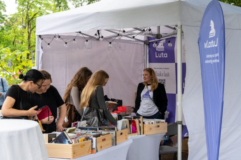 Balta telts ar Luta grāmatu stendu, kurā sievietes aplūko grāmatas koka kastēs uz galdiem. Labajā pusē sieviete baltā t-kreklā ar uzrakstu "Luto.lv" un melnā žaketē stāv pie stenda, smaidot un skatoties pa kreisi. Uz purpursarkaniem baneriem redzams uzraksts "Luta", "Pievienojies un atbalsti" un "Laiks lasīt".