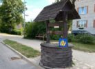 Koka aka ar jumtiņu stāv uz bruģētas ietves blakus ielai. Uz akas ir zila zīme ar dzeltenām stilizētām figūrām un uzrakstu "ANF". Fonā redzama gaišu ķieģeļu ēka ar melnu automašīnu priekšā un koku rinda gar ielu. Aiz akas ir ielas zīme ar tekstu "Pasta iela".