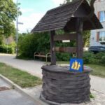 Koka aka ar jumtiņu stāv uz bruģētas ietves blakus ielai. Uz akas ir zila zīme ar dzeltenām stilizētām figūrām un uzrakstu "ANF". Fonā redzama gaišu ķieģeļu ēka ar melnu automašīnu priekšā un koku rinda gar ielu. Aiz akas ir ielas zīme ar tekstu "Pasta iela".