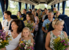 Autobusā sēž dažādu paaudžu cilvēku grupa, kuri smaida un tur rokās krāsainus pļavas ziedu pušķus. Aiz loga redzama zaļa ainava.