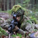 Karavīrs kamuflāžas tērpā ar sejas maskējumu un ķiveri, kas rotāta ar sūnām un zāli, guļ mežā un tēmē ar šauteni ar sarkano punktu tēmēkli.