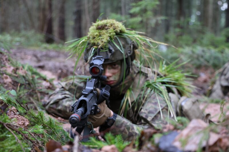 Karavīrs kamuflāžas tērpā ar sejas maskējumu un ķiveri, kas rotāta ar sūnām un zāli, guļ mežā un tēmē ar šauteni ar sarkano punktu tēmēkli.