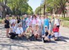 Pieaugušo grupa, pārsvarā sievietes, smaidot pozē saulainā dienā uz koku ieskautas pilsētas promenādes. Viena sieviete labajā pusē tur karti vai planšeti.