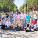 Pieaugušo grupa, pārsvarā sievietes, smaidot pozē saulainā dienā uz koku ieskautas pilsētas promenādes. Viena sieviete labajā pusē tur karti vai planšeti.