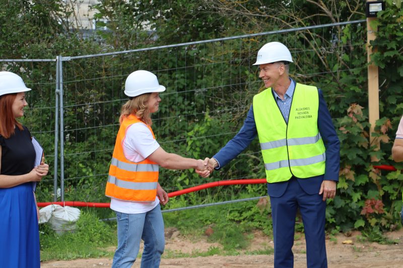 Divi vīrieši, viens oranžā un otrs dzeltenā vestē, smaidot paspiež viens otram roku. 