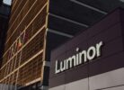 Luminor ēka ar uzrakstu "Luminor", kuras priekšā mastos plīvo Latvijas, Lietuvas un Ukrainas karogi. Ēku apspīd silta, zeltaina gaisma.