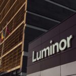 Luminor ēka ar uzrakstu "Luminor", kuras priekšā mastos plīvo Latvijas, Lietuvas un Ukrainas karogi. Ēku apspīd silta, zeltaina gaisma.