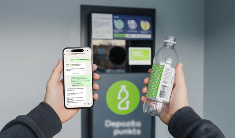 Personas rokas tuvplānā tur viedtālruni un tukšu plastmasas pudeli ar zaļu etiķeti. Viedtālruņa ekrānā redzams ziņojumapmaiņas logs ar virsrakstu "Depozīta punkta asistents". Sarunā jautāts "Sveiki! Kur var nodot iepakojumu?" un atbilde ir "Es esmu Depozīta punkts virtuālais asistents." ar iespējām, piemēram, "Tuvākais taromāts". Fonā atrodas depozīta automāts ar zaļu pudeles simbola logotipu un uzrakstu "Depozīta punkts".
