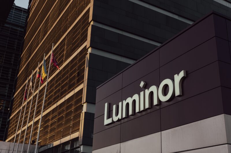 Luminor ēka ar uzrakstu "Luminor", kuras priekšā mastos plīvo Latvijas, Lietuvas un Ukrainas karogi. Ēku apspīd silta, zeltaina gaisma.