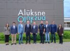 Deviņi cilvēki, tostarp septiņi vīrieši un divas sievietes, stāv rindā, pozējot pie modernas ēkas, uz kuras fasādes redzams liels uzraksts "Alūksne Oluksna".