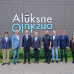 Deviņi cilvēki, tostarp septiņi vīrieši un divas sievietes, stāv rindā, pozējot pie modernas ēkas, uz kuras fasādes redzams liels uzraksts "Alūksne Oluksna".