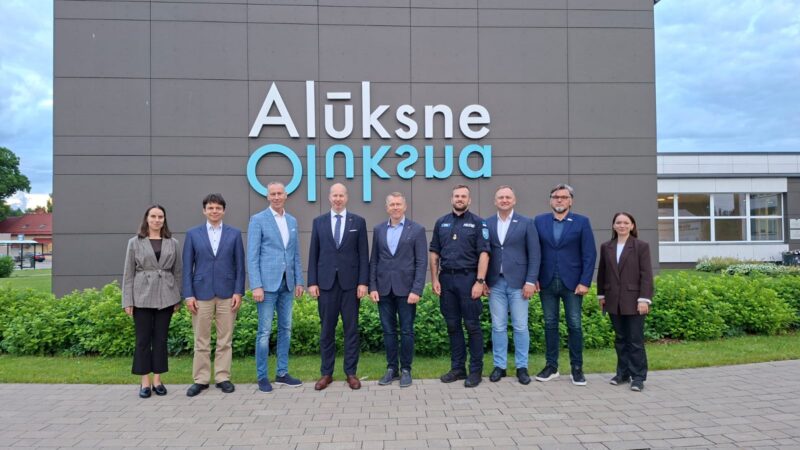 Deviņi cilvēki, septiņi vīrieši un divas sievietes, stāv rindā pie modernas ēkas, uz kuras redzams liels zils uzraksts "Alūksne Oluksna".