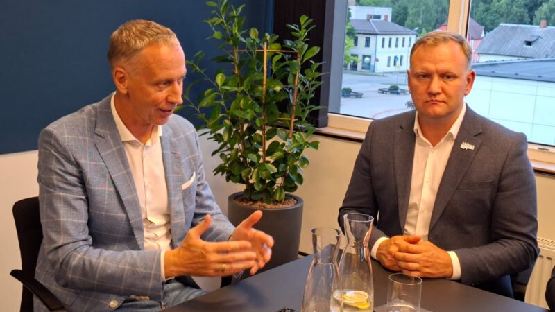 Divi vīrieši sēž pie galda. Vīrietis kreisajā pusē, gaiši zilā rūtota žaketē, runā un žestikulē. Vīrietis labajā pusē, tumšā žaketē, skatās uz priekšu, salicis rokas uz galda. Starp abiem ir zaļš telpaugs, un fonā pa logu redzamas ēkas.