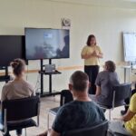 Sieviete dzeltenā kreklā vada prezentāciju sēdošai cilvēku grupai. Lielajā ekrānā aiz viņas redzams teksts "Veselīgs uzturs" un "Veselīgs dzīvesveids". Blakus ir vēl viens tukšs ekrāns un tāfele.