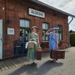 Vīrietis melnā kreklā un platmalē pasniedz brūnu koferi zēnam, kurš kāpj melnā tvaika lokomotīvē. Uz lokomotīves sāniem redzami uzraksti "Ferdinand" un "BĀNĪŠA ZEME". Aiz lokomotīves kabīnes ir malkas tenderis, aiz kura redzama sievietes galva.