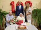 Vecāka sieviete svin 100. dzimšanas dienu, sēžot pie galda ar ziediem un torti, kas dekorēta ar ogām un skaitli "100". Blakus sēž vīrietis ratiņkrēslā, bet aizmugurē stāv vīrietis uzvalkā un sieviete. Fonā redzami sarkani baloni "100". Uz galda ir maisiņš ar uzrakstu "Alūksne".