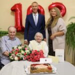 Vecāka sieviete svin 100. dzimšanas dienu, sēžot pie galda ar ziediem un torti, kas dekorēta ar ogām un skaitli "100". Blakus sēž vīrietis ratiņkrēslā, bet aizmugurē stāv vīrietis uzvalkā un sieviete. Fonā redzami sarkani baloni "100". Uz galda ir maisiņš ar uzrakstu "Alūksne".