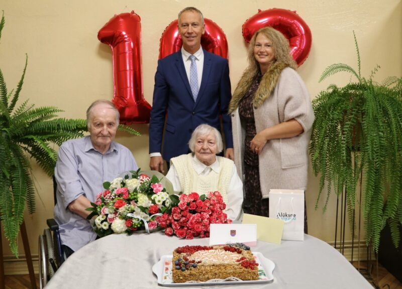 Vecāka sieviete svin 100. dzimšanas dienu, sēžot pie galda ar ziediem un torti, kas dekorēta ar ogām un skaitli "100". Blakus sēž vīrietis ratiņkrēslā, bet aizmugurē stāv vīrietis uzvalkā un sieviete. Fonā redzami sarkani baloni "100". Uz galda ir maisiņš ar uzrakstu "Alūksne".