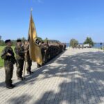 Karavīri formastērpos stāv ierindā uz bruģēta laukuma Daži karavīri tur šautenes. Fonā zaļš zāliens, koki, ūdenskrātuve un zilas debesis.