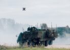 Tumšs militārais drons lido virs zaļa bruņutransportiera, kas pārklāts ar maskēšanās tīklu un atrodas uz grants ceļa. Transportlīdzekli apņem dūmi vai putekļi, fonā redzamas citas militārās mašīnas un mežs.