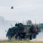 Tumšs militārais drons lido virs zaļa bruņutransportiera, kas pārklāts ar maskēšanās tīklu un atrodas uz grants ceļa. Transportlīdzekli apņem dūmi vai putekļi, fonā redzamas citas militārās mašīnas un mežs.