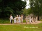 Grupa cilvēku tradicionālos tērpos, daži ar siena grābekļiem un dakšām, dodas pa zemes ceļu gar koka ēkām un zaļumiem. Attēlā redzams uzraksts: Pļaujas svētkiem 40.