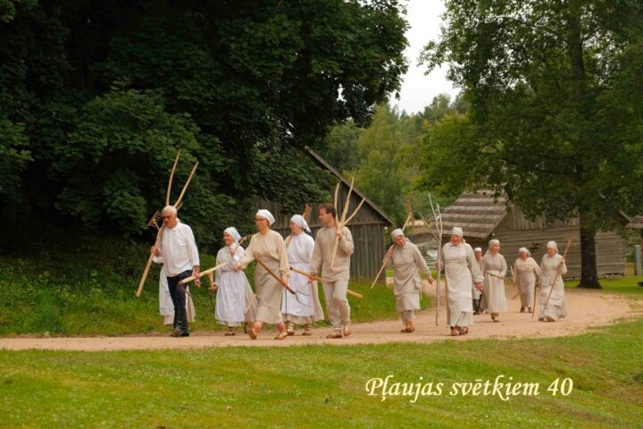 Grupa cilvēku tradicionālos tērpos, daži ar siena grābekļiem un dakšām, dodas pa zemes ceļu gar koka ēkām un zaļumiem. Attēlā redzams uzraksts: Pļaujas svētkiem 40.