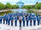 Militārais orķestris ar aptuveni 50 dalībniekiem zilās uniformās stāv ārā, turot mūzikas instrumentus. Fonā redzams dīķis un liela, moderna stikla ēka.