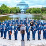 Militārais orķestris ar aptuveni 50 dalībniekiem zilās uniformās stāv ārā, turot mūzikas instrumentus. Fonā redzams dīķis un liela, moderna stikla ēka.