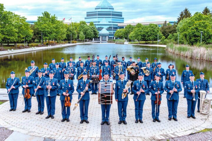 Militārais orķestris ar aptuveni 50 dalībniekiem zilās uniformās stāv ārā, turot mūzikas instrumentus. Fonā redzams dīķis un liela, moderna stikla ēka.