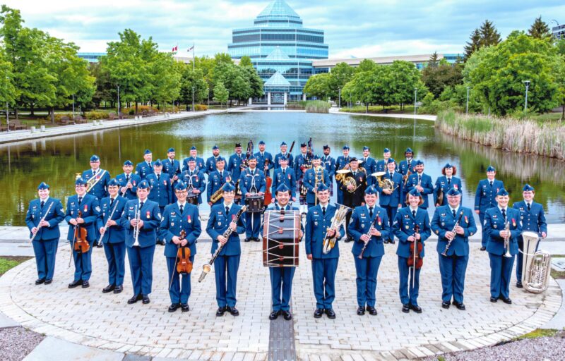 Militārais orķestris ar aptuveni 50 dalībniekiem zilās uniformās stāv ārā, turot mūzikas instrumentus. Fonā redzams dīķis un liela, moderna stikla ēka.