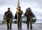 Trīs karavīri kamuflāžas uniformās un melnās beretēs stāv ārā. Divi no viņiem tur šautenes, bet vidējais karavīrs tur dzeltenu karogu ar zilu apmali. Fonā redzama ūdenskrātuve, koki un gaišs pikaps.