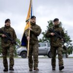 Trīs karavīri kamuflāžas uniformās un melnās beretēs stāv ārā. Divi no viņiem tur šautenes, bet vidējais karavīrs tur dzeltenu karogu ar zilu apmali. Fonā redzama ūdenskrātuve, koki un gaišs pikaps.