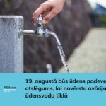 Roka atver ūdens krānu uz akmens pīlāra, no kura tek ūdens. Attēlā redzams teksts: Alūksne Novads un 19. augustā būs ūdens padeves atslēgums, lai novērstu avāriju ūdensvada tīklā.