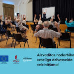 Zālē vecāka gadagājuma cilvēki sēž uz krēsliem un veic vingrojumus ar paceltām rokām, sekojot instruktoram priekšā. Attēlā redzams teksts: "Aizvadītas nodarbības veselīga dzīvesveida veicināšanai", un logotipi "Līdzfinansē Eiropas Savienība", "2027 Nacionālais attīstības plāns", "Alūksne".
