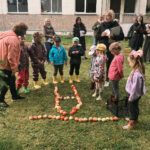 Pieaugušo grupa stāv uz zāles laukuma netālu no asfaltēta celiņa, skatoties uz lielu bērzu un rotaļu laukumu. Pa labi redzama ēka un mākoņainas rudens debesis.