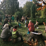 Pieaugušo grupa stāv zāliena rotaļu laukumā pie ēkas, vērsta pret lielu koku zem mākoņainām debesīm.